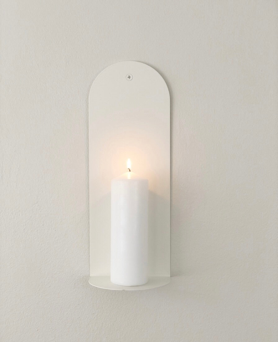 Wall-mounted candle holder with a lit candle on a plain wall. Avaiable in white, grey, stone, black. The Bold wall sconce in Bone White mounted on textured white wall, Nomad Studio.
