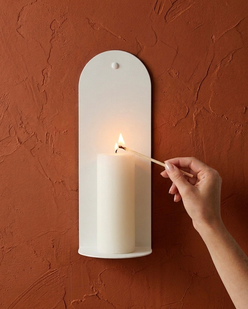 Hand lighting a pillar candle on a white wall sconce against a textured red-brown wall. Bone White Bold wall sconce on terracotta wall with hand lighting candle, Nomad Studio.